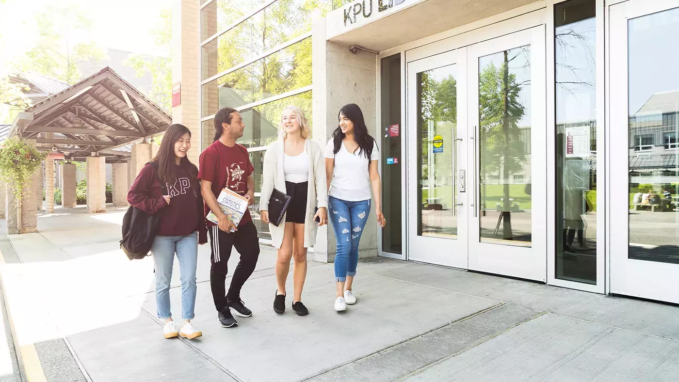 Image of four KPU students outside the KPU Surrey library. They are smiling as they walk and chat with each other.